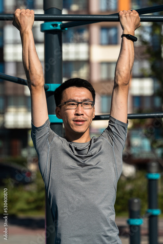 Adult asian man in glasses drinking working out on exercise equipment in park.
