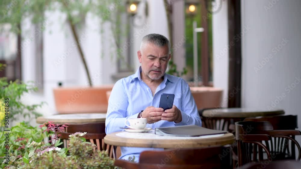 Handsome caucasian middle aged businessman sitting at the street cafe uses phone texting scrolling tapping. Man sitting at table in street cafe enjoying morning coffee. Handsome caucasian middle aged businessman sitting at the street cafe uses phone texting scrolling tapping. Man sitting at table in street cafe enjoying morning coffee.