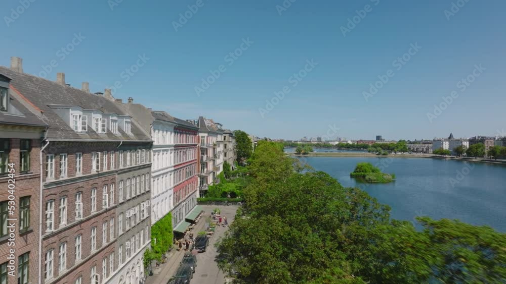 Forwards fly above lush green trees on lake bank. Row of townhouses on waterfront in city. Sunny day with clear blue sky. Copenhagen, Denmark Forwards fly above lush green trees on lake bank. Row of townhouses on waterfront in city. Sunny day with clear blue sky. Copenhagen, Denmark