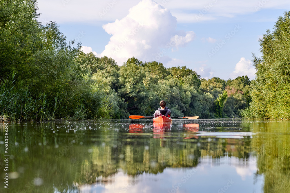 Obraz premium Rear view of man in red kayak. Kayaking on river near the shore with green trees against the blue sky and clouds in the background