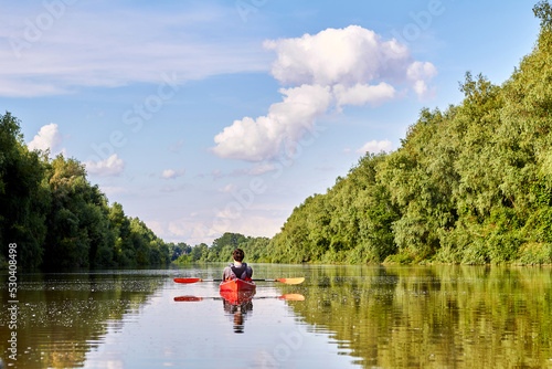 Rear view of man in red kayak. Kayaking on river near the shore with green trees against the blue sky and clouds in the background