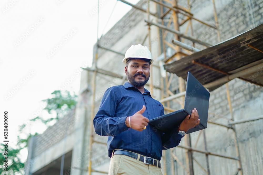 young Indian engineer in uniform and using laptop at field
