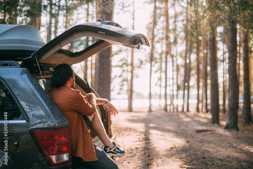 A young man is sitting relaxed alone in the open trunk of a car in the woods by the lake at sunset. photo