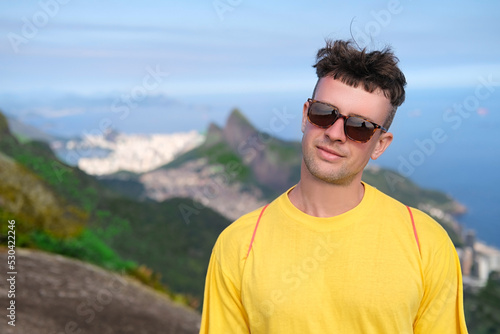 Handsome young man in sunglasses smiles and stands on a mountain against the backdrop of mountains and ocean