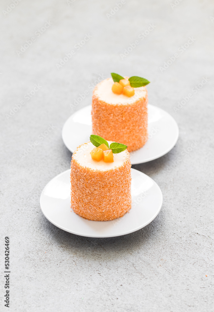 Vegan dessert. Apricot mousse cream chocolate dessert of cylindrical shape, sprinkled with colored coconut flakes, on a plate. Light gray background Vegan dessert. Apricot mousse cream chocolate dessert of cylindrical shape, sprinkled with colored coconut flakes, on a plate. Light gray background