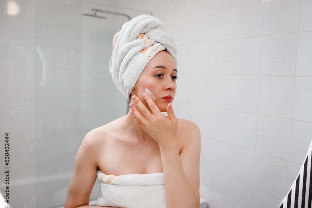 Young woman applying anti-wrinkle cream standing behind mirror in home bathroom. Cosmetology and beauty procedure. Skin care after cleansing Young woman applying anti-wrinkle cream standing behind mirror in home bathroom. Cosmetology and beauty procedure. Skin care after cleansing