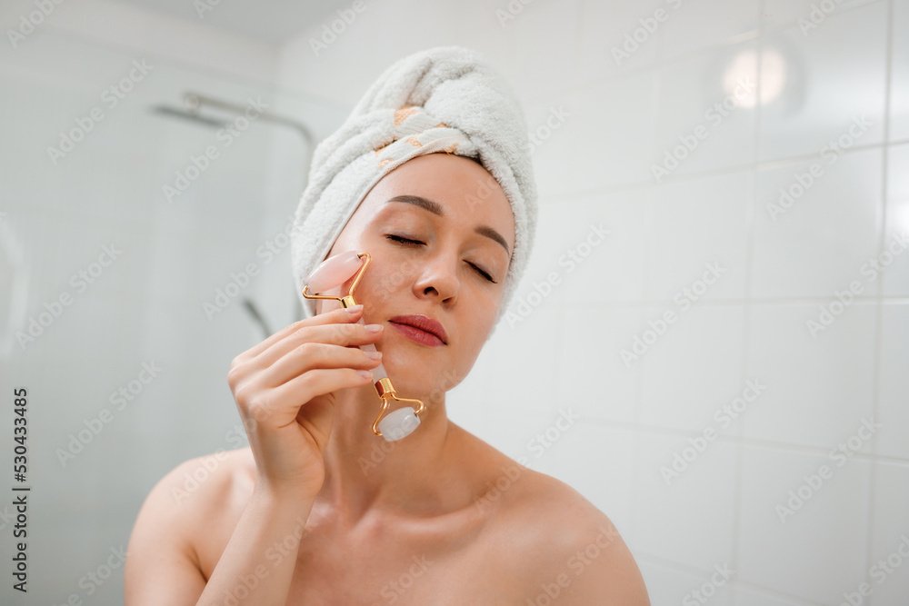 Beautiful woman with perfect skin wearing white towel after shower, making face massage using a jade face roller with natural quartz stone in bathroom. Natural cosmetics concept, wrinkle smoothing Beautiful woman with perfect skin wearing white towel after shower, making face massage using a jade face roller with natural quartz stone in bathroom. Natural cosmetics concept, wrinkle smoothing