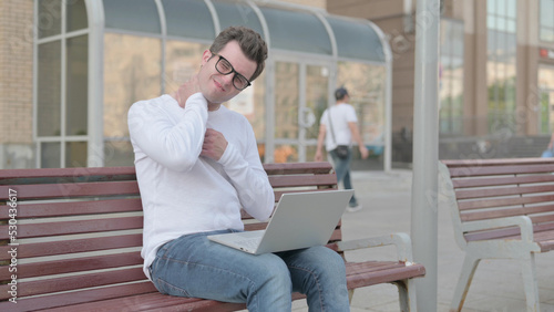 Young Man with Neck Pain Using Laptop while Sitting Outdoor on Bench