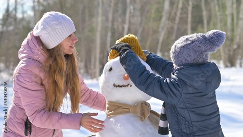 Winter fun. Happy mom and son are building a snowman in a snowy park, putting on scarf and hat. Lovely family at winter holidays.