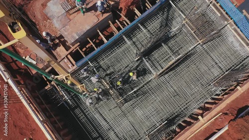 Drone sobrevoando um prédio sendo construído. Homens ao longe trabalhando na concretagem das fundações, jogando cimento e espalhando. Ferros de construção instalados. photo