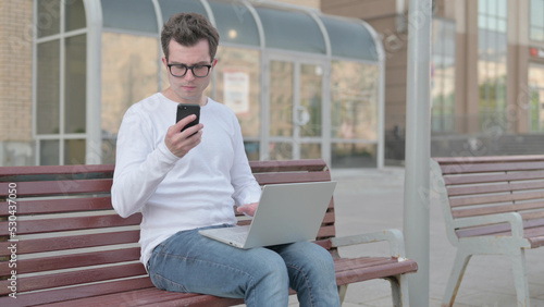 Young Man Using Smartphone and Laptop while Sitting Outdoor on Bench