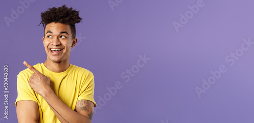 Close-up portrait of funny cute hispanic man with dreads, laughing and smiling at something, pointing looking upper left corner enthusiastic expression, purple background photo