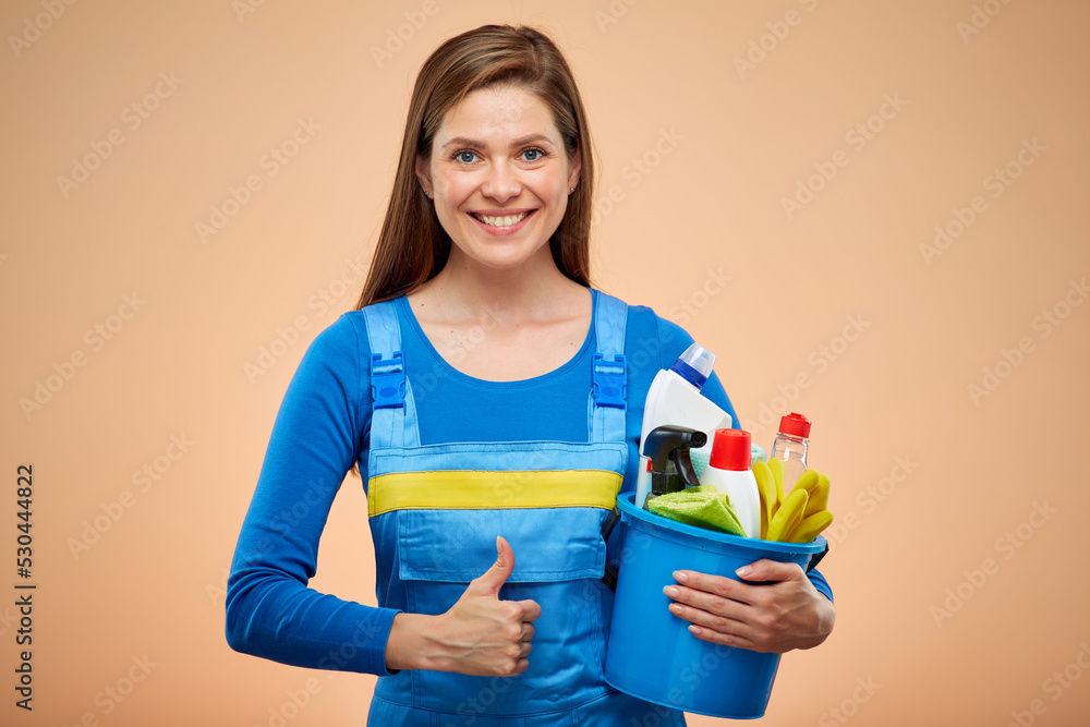 Woman cleaner in overalls holding cleaning products in bucket. isolated female housekeeper portrait on brown beige background. Woman cleaner in overalls holding cleaning products in bucket. isolated female housekeeper portrait on brown beige background.