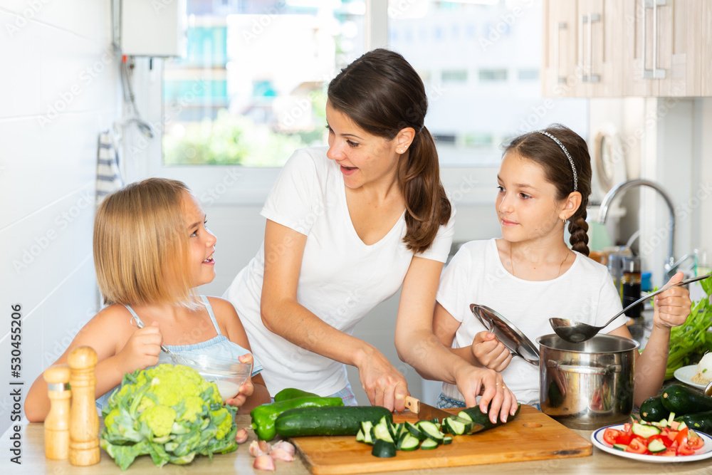Cheerful woman with two little daughters prepare vegetable salad in the kitchen at home