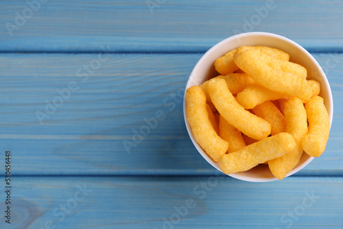 Bowl of tasty cheesy corn puffs on blue wooden table, top view. Space for text