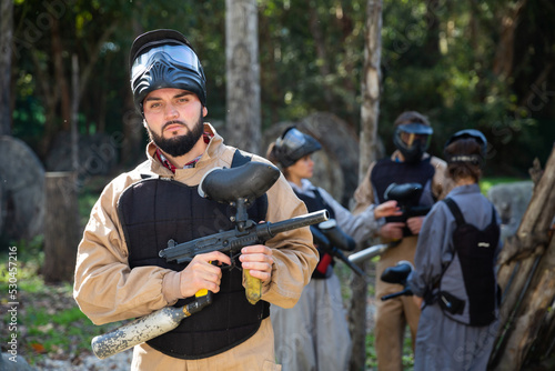 Portrait of male paintball sport player holding shooting gun and his team on background