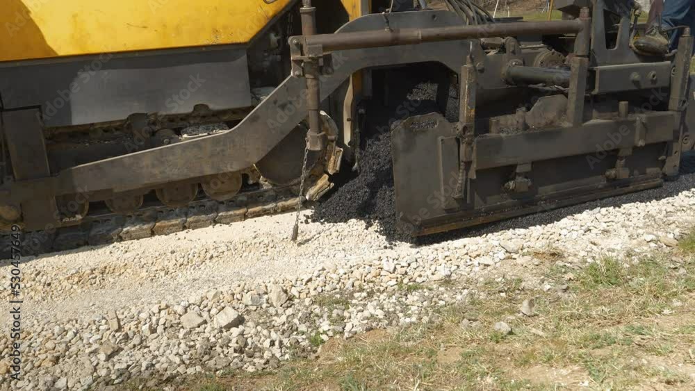 CLOSE UP: Asphalt paver machine dumping and spreading blacktop over crushed rock. Construction workers operating with heavy machinery for laying black asphalt surface at the driveway in morning light. CLOSE UP: Asphalt paver machine dumping and spreading blacktop over crushed rock. Construction workers operating with heavy machinery for laying black asphalt surface at the driveway in morning light.