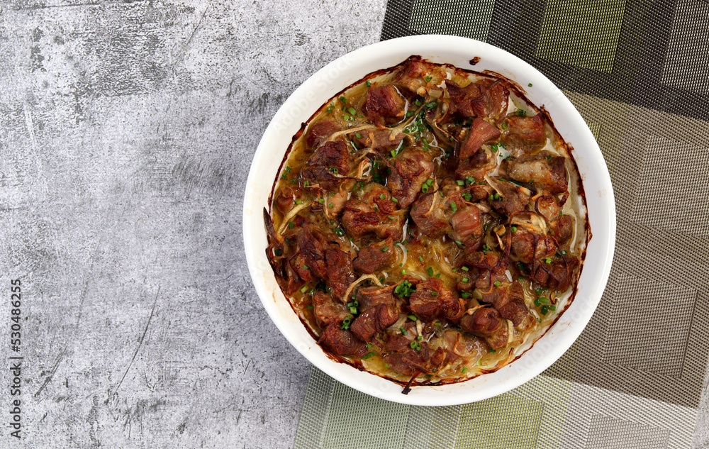 Braised pork with onions in a white baking dish on a dark grey background. Top view, flat lay Braised pork with onions in a white baking dish on a dark grey background. Top view, flat lay