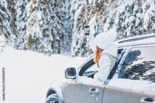 Teenager girl traveling looking out of car window and taking selfie on smartphone in winter snowy forest. Road trip and local travel concept. Happy child enjoying car ride.