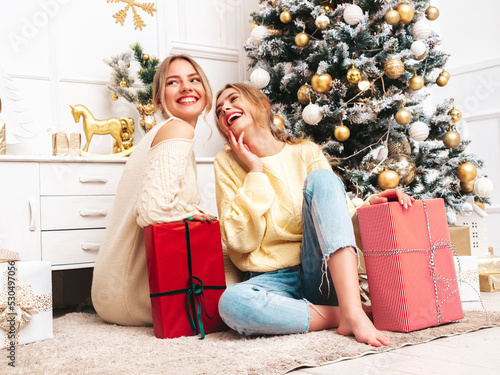 Two beautiful blond women. Models posing near decorated Christmas tree at New Year eve. Having fun, ready for celebration. Bright holiday of best friends dressed in warm winter sweaters photo