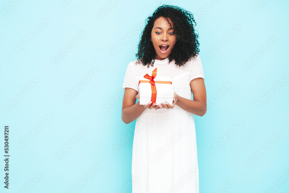 Beautiful black woman with afro curls hairstyle. Smiling model dressed in white summer dress. female posing near blue wall in studio. Tanned and cheerful. Holding gift box. Shocked, surprised Beautiful black woman with afro curls hairstyle. Smiling model dressed in white summer dress. female posing near blue wall in studio. Tanned and cheerful. Holding gift box. Shocked, surprised
