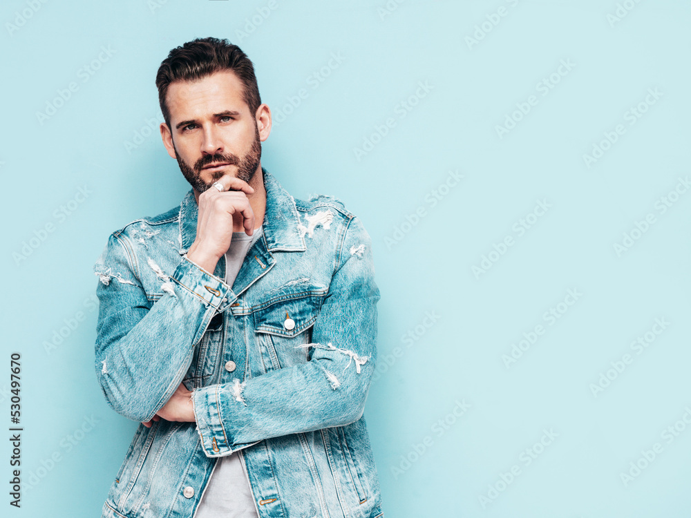 Portrait of handsome confident stylish hipster lambersexual model. Sexy man dressed in jeans jacket. Fashion male isolated on blue wall in studio Portrait of handsome confident stylish hipster lambersexual model. Sexy man dressed in jeans jacket. Fashion male isolated on blue wall in studio