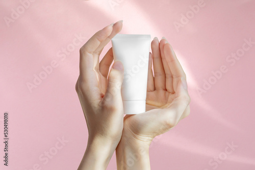 Woman's hands are holding an unmarked plastic container for cosmetics. White bottle for cream, toiletries on pink isolated background. Concept of skin care. Close-up, mockup style