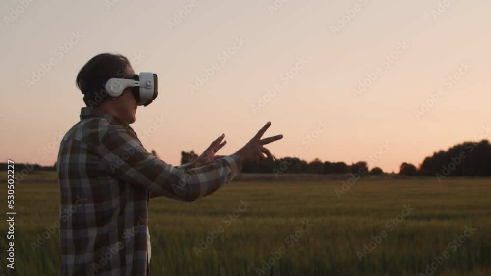 Farmer in virtual reality helmet in front of a sunset agricultural landscape. Man in a countryside field. Country life, food production, farming and technology concept . Farmer in virtual reality helmet in front of a sunset agricultural landscape. Man in a countryside field. Country life, food production, farming and technology concept .