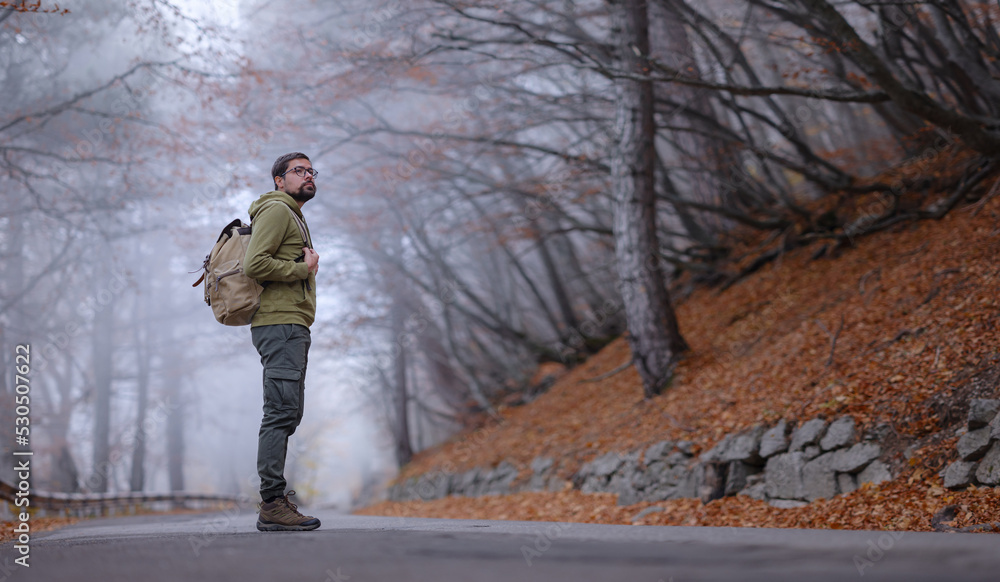 Young handsome man posing in autumn forest on road. young hipster guy with backpack , traveller standing in woods, Hiking, Forest, Journey, active healthy lifestyle, adventure, vacation concept.