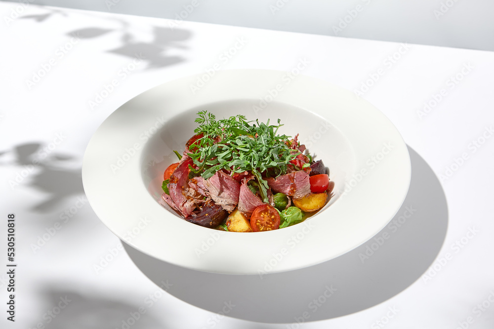 Meat salad with pastrami, baked vegetables and arugula on white background with hard shadows. Salat with roasted beef and vegetables on summer lunch with shadows of leaves. Meat salad with pastrami, baked vegetables and arugula on white background with hard shadows. Salat with roasted beef and vegetables on summer lunch with shadows of leaves.