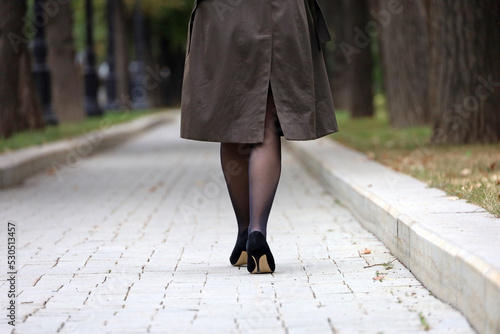 Woman in raincoat, black stockings and shoes on high heels walking in city park, rear view. Female fashion in autumn