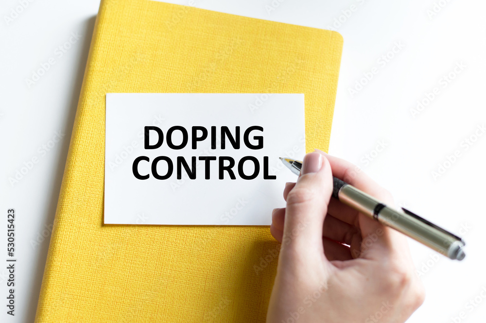 DOPING CONTROL text on a card on the background of a notebook on a table next to a person's hand with a pen DOPING CONTROL text on a card on the background of a notebook on a table next to a person's hand with a pen