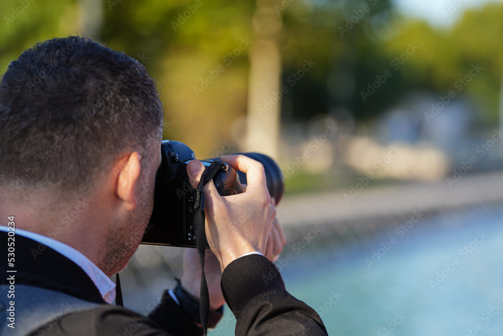 Male photographer with dark gray hair photographed from behind while is taking pictures with his DSLR photo camera during a holiday. Photography as a hobby. Male photographer with dark gray hair photographed from behind while is taking pictures with his DSLR photo camera during a holiday. Photography as a hobby.