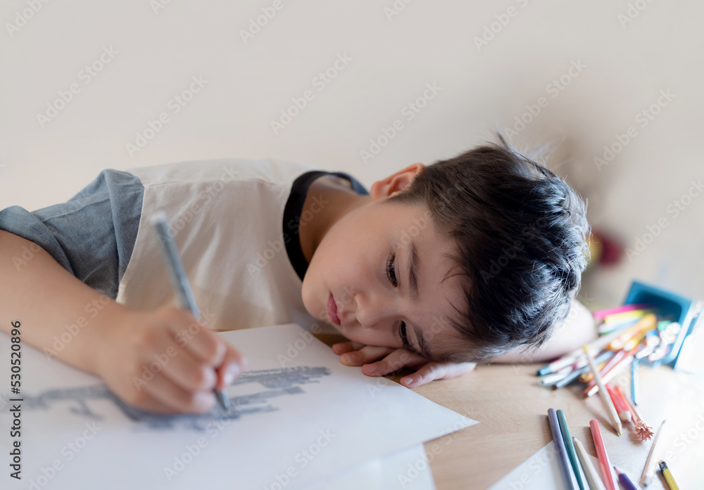 Kid drawing on paper, Portrait Boy lying head down on hand while using grey pen painting, Child with thinking face doing art and craft for homework. Kid drawing on paper, Portrait Boy lying head down on hand while using grey pen painting, Child with thinking face doing art and craft for homework.