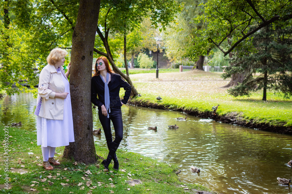 Fototapeta premium Two women together - blonde in light and redhead in dark clothes posing in a park. Cosplay - female version of Angel and Demon (Good Omens)