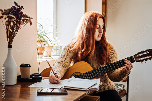 Unaltered candid portrait of young red haired woman in sweater playing acoustic guitar sitting by window at home. Hobbies, indoor home activities for adults in winter, autumn.