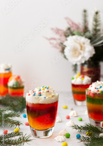 Glasses with multi-colored jelly, decorated with cream and dragees, meringues and marshmallows, Turkish delight and marmalade, drinks among the fir branches on the holiday table