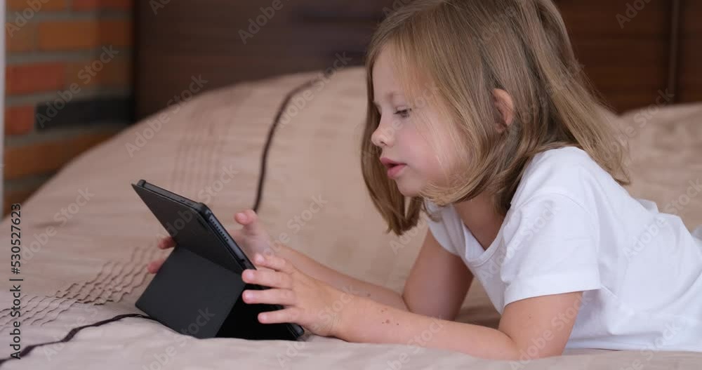 Beautiful charming caucasian little girl talking playing with tablet at home on bed Beautiful charming caucasian little girl talking playing with tablet at home on bed