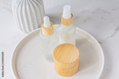 Skincare products set. Natural moisturizer cream jar, dropper bottle with serum and essence on round ceramic tray on marble background. Organic cosmetics concept