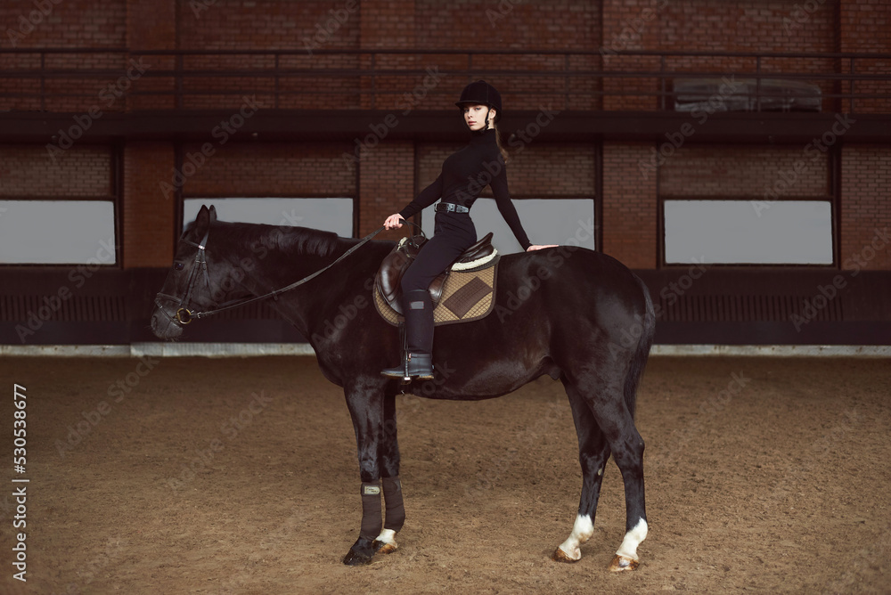 Young beautiful woman siting horseback in dark indoor manege Woman jockey in a black uniform with his black horse. Arena horse. Equestrian sport. Racecourse. Sport and fashion Young beautiful woman siting horseback in dark indoor manege Woman jockey in a black uniform with his black horse. Arena horse. Equestrian sport. Racecourse. Sport and fashion
