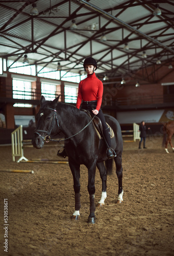 Young beautiful woman in red doing horseback inside dark manege. Jockey during training and dressage. Sport concept.
