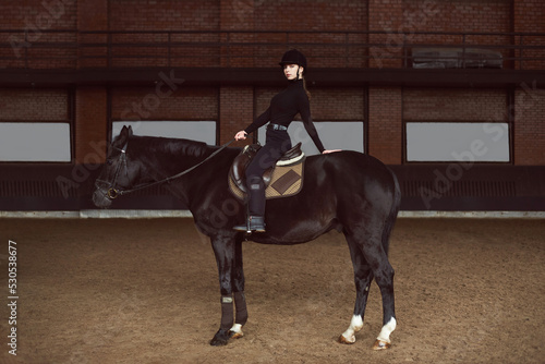 Young beautiful woman siting horseback in dark indoor manege Woman jockey in a black uniform with his black horse. Arena horse. Equestrian sport. Racecourse. Sport and fashion  © Oksana