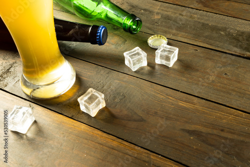 Image of beer bottles, ice cubes and pint glass of beer on wooden table, with copy space