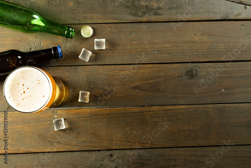 Image of two glass beer bottles, pint of beer and ice cubes lying on wooden table, with copy space