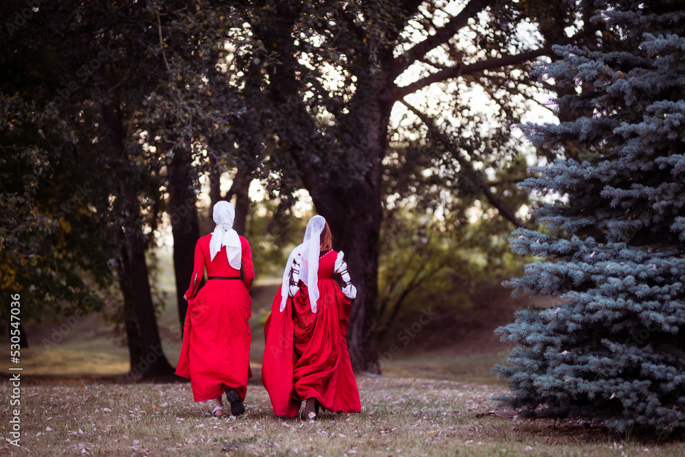 Living history: a 15th-century woman dressed as a wealthy lady in a Venice dress and her maid in the woods, back view. Living history: a 15th-century woman dressed as a wealthy lady in a Venice dress and her maid in the woods, back view.
