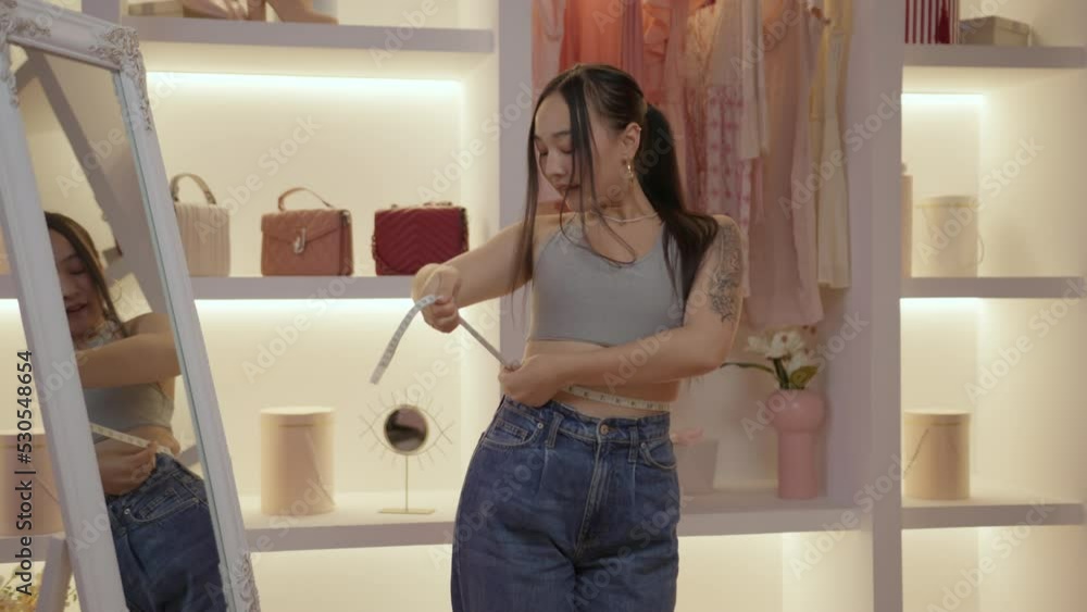 Positive young Asian woman measuring her waist with tape measure looking at mirror. Slim woman is happy with weight loss. Positive young Asian woman measuring her waist with tape measure looking at mirror. Slim woman is happy with weight loss.