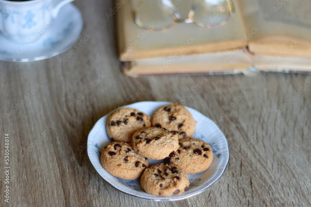 Cup of tea, plate with chocolate chip cookies, open book, reading glasses, lit candles and dry lavender flowers. Hygge at home, selective focus.