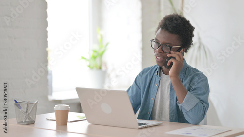 Young African Man Talking on Phone while using Laptop in Office
