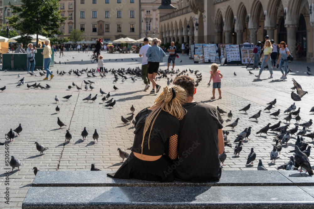 Obraz premium Couple sitting on a marble bench outdoors in the city square on a sunny summer day, traditional young couple, tourists in Krakow, close relationship, love