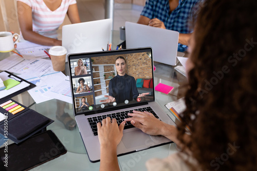 Biracial businesswoman at desk in office making laptop video call with diverse colleagues on screen